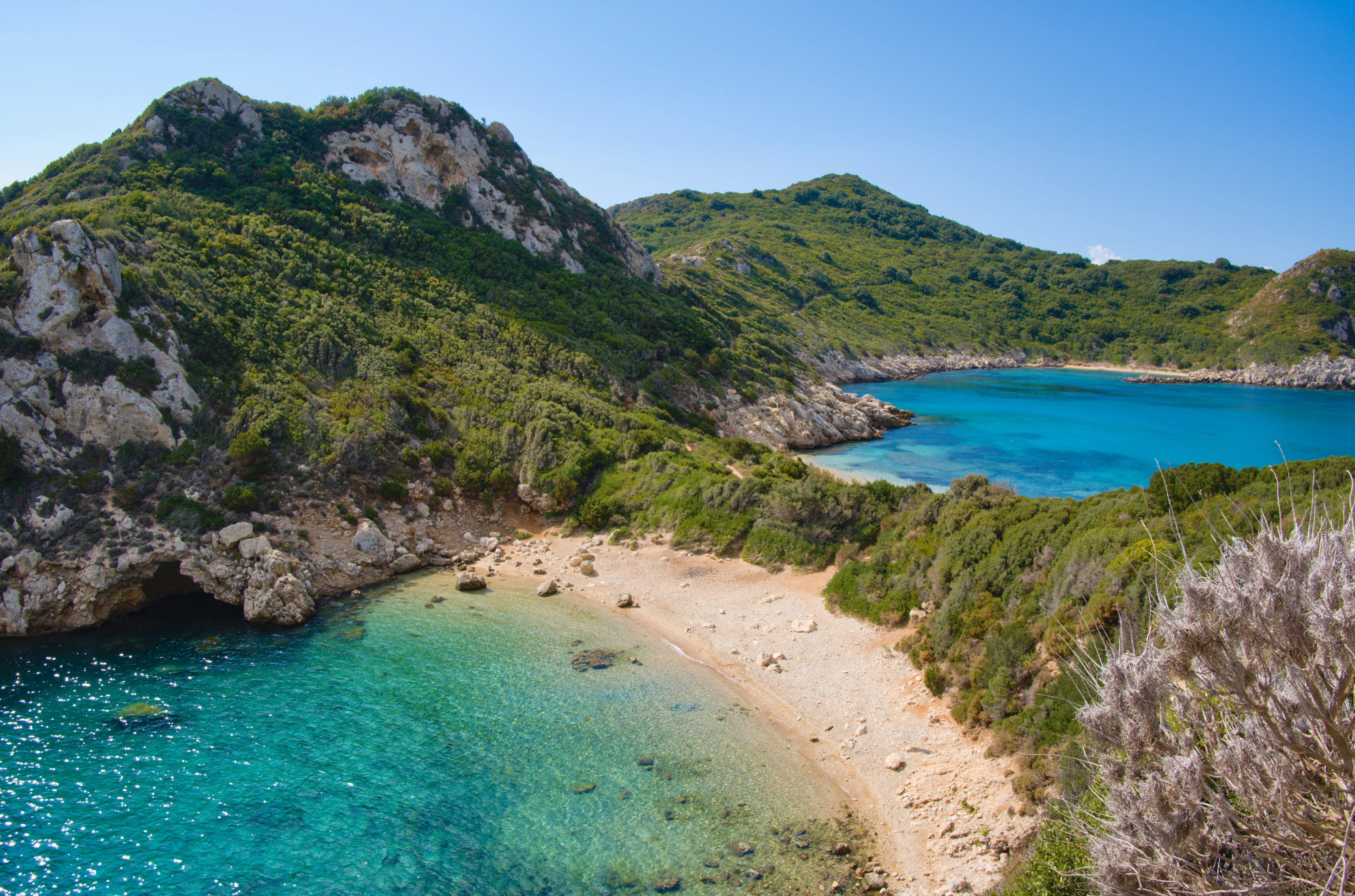 The twin bays of Porto Timoni on Corfu's west coast seen from the headland path, a pale pebble isthmus dividing two coves of clear turquoise water below scrub-covered cliffs.