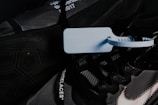 A close-up image of a dark-colored sneaker featuring a prominent blue zip tie attached to the laces. The shoe has text on the laces and a visible, stylized Nike logo with a stitch pattern. Details of the outsole and design elements are visible, with a hexagonal pattern on the tread.