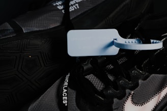 A close-up image of a dark-colored sneaker featuring a prominent blue zip tie attached to the laces. The shoe has text on the laces and a visible, stylized Nike logo with a stitch pattern. Details of the outsole and design elements are visible, with a hexagonal pattern on the tread.