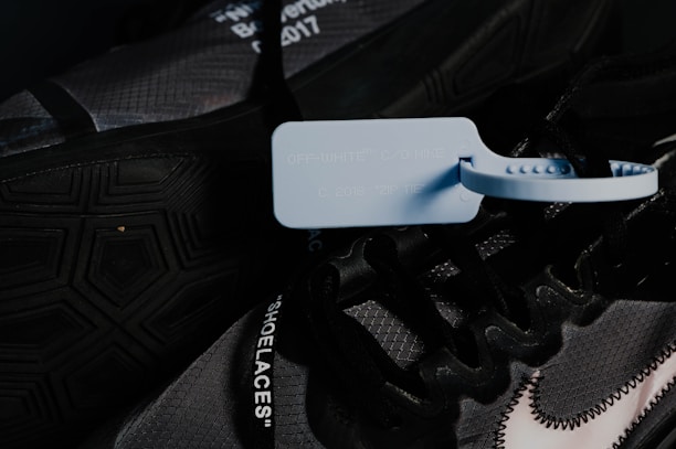 A close-up image of a dark-colored sneaker featuring a prominent blue zip tie attached to the laces. The shoe has text on the laces and a visible, stylized Nike logo with a stitch pattern. Details of the outsole and design elements are visible, with a hexagonal pattern on the tread.