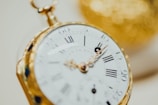 A close-up view of a vintage pocket watch with a gold casing. The watch face displays Roman numerals and intricate detailing, with the focus on the minute and hour hands.
