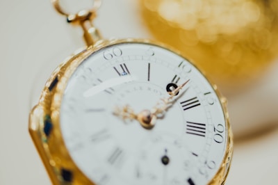 A close-up view of a vintage pocket watch with a gold casing. The watch face displays Roman numerals and intricate detailing, with the focus on the minute and hour hands.
