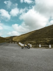 A solitary motorcycle is parked in an empty lot surrounded by a vast, open landscape. Rolling hills covered in green grass stretch out under a partly cloudy blue sky. The scene evokes a sense of freedom and tranquility.