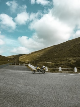 A solitary motorcycle is parked in an empty lot surrounded by a vast, open landscape. Rolling hills covered in green grass stretch out under a partly cloudy blue sky. The scene evokes a sense of freedom and tranquility.