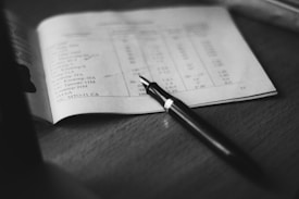 A black and white photograph depicting a close-up view of a fountain pen resting on an open notebook. The notebook displays text and numbers in a tabular format. The background includes a smooth surface, possibly a desk or table.