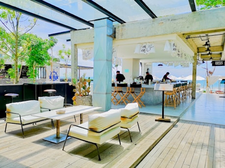 A modern outdoor cafe with white cushioned seating and wooden decking, featuring a bar area with high stools. The space is decorated with minimalistic Japanese elements such as hanging banners with Japanese characters. Green trees and a view of the ocean are visible in the background, enhancing the open-air and relaxed atmosphere.