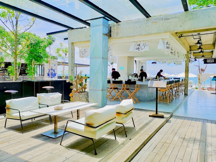 A modern outdoor cafe with white cushioned seating and wooden decking, featuring a bar area with high stools. The space is decorated with minimalistic Japanese elements such as hanging banners with Japanese characters. Green trees and a view of the ocean are visible in the background, enhancing the open-air and relaxed atmosphere.