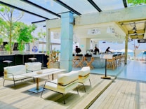 A modern outdoor cafe with white cushioned seating and wooden decking, featuring a bar area with high stools. The space is decorated with minimalistic Japanese elements such as hanging banners with Japanese characters. Green trees and a view of the ocean are visible in the background, enhancing the open-air and relaxed atmosphere.