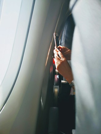 A person is holding a smartphone near an airplane window. Sunlight is streaming in, creating a bright and serene atmosphere inside the cabin. The interior of the plane, including seats and window, is visible. The person's wristwatch can also be seen.