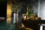 A modern indoor setting features a reflective surface with a potted plant arrangement, including desert plants and cacti, against a textured glass backdrop. A ceramic vase with green foliage stands on the left, contributing to the natural aesthetic. The scene is softly lit, emphasizing the greenery and creating a serene atmosphere.