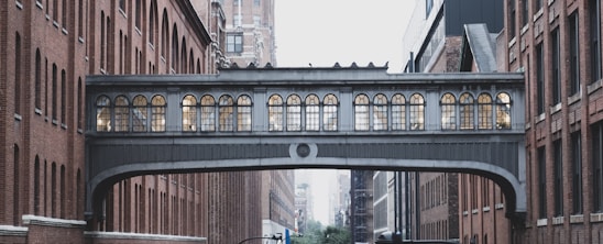 A bridge with arched windows connects two historic brick buildings in an urban setting. The architecture features intricate design elements and the surrounding area is densely built with similar structures.