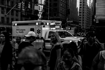 A busy urban street scene with a crowd of people walking past an emergency medical service ambulance. Tall buildings surround the area, and various signs are visible in the background.