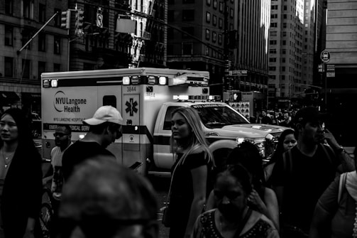 A busy urban street scene with a crowd of people walking past an emergency medical service ambulance. Tall buildings surround the area, and various signs are visible in the background.