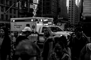 A busy urban street scene with a crowd of people walking past an emergency medical service ambulance. Tall buildings surround the area, and various signs are visible in the background.