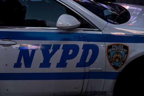 A close-up view of a New York Police Department (NYPD) vehicle with a visible blue stripe and a police department badge on the door. The vehicle's white paint contrasts with the blue lettering.