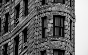 A detailed close-up of a building's architectural facade featuring ornate geometric patterns and decorative elements in a grayscale color scheme. Large windows and air conditioning units are visible among the intricate stonework.