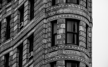 A detailed close-up of a building's architectural facade featuring ornate geometric patterns and decorative elements in a grayscale color scheme. Large windows and air conditioning units are visible among the intricate stonework.