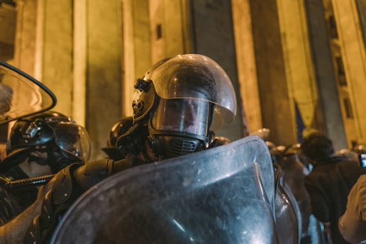 Several uniformed officers wearing riot gear, including helmets and face shields, stand closely together against a backdrop of tall, illuminated stone structures, suggesting an urban or monumental setting.