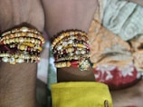 Several colorful bracelets with beads, pearls, and intricate designs adorn the wrists of two individuals. The bracelets feature a mix of gold, red, and pearl accents, with one prominently featuring a floral motif embellished with gemstones.