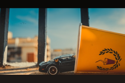 A toy car styled after a classic muscle car is partially covered by a large yellow cup with a laurel wreath design. The scene appears to be set outdoors and blurred buildings can be seen in the background, along with a metal railing and a clear sky.