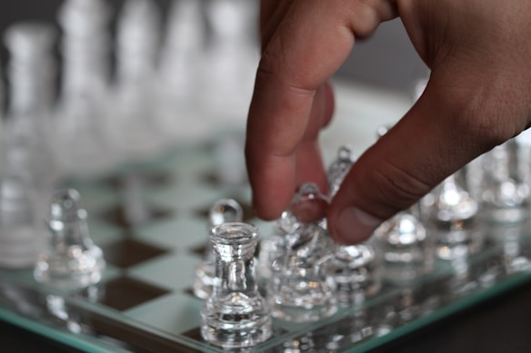 A hand is seen picking up a transparent chess piece from a glass chessboard. Other chess pieces are also visible on the board, made of the same transparent material. The background is blurred, keeping the focus on the chessboard and the hand.