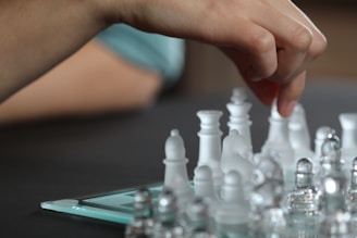 A hand is reaching over a chessboard, gently holding a chess piece. The chess pieces are made of translucent glass, standing among white pieces, and there is a digital device partially visible beneath the board. The background is softly blurred, drawing focus to the hand and the chess set.