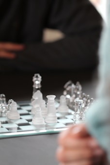 A close-up of a glass chess board with both clear and frosted pieces arranged in various positions. In the background, a person dressed in dark clothing sits with their arms crossed, watching or pondering the game. The focus is on the chess board and pieces, creating a sense of strategy and concentration.