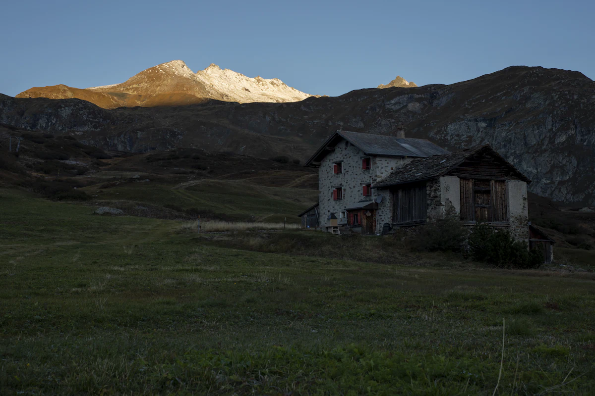 An old stone farmhouse in the meadows at Bivio, in shadow below a sunlit snowy peak