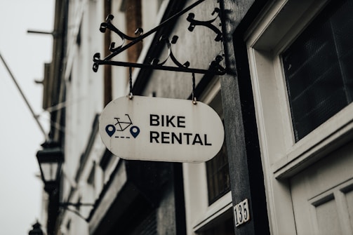 A metal sign with the words 'Bike Rental' hangs from a decorative bracket on a building. The sign includes icons of a bicycle and navigation symbols. The building has a white door and the number '135' visible. The atmosphere appears overcast, contributing to a muted tone.