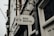 A metal sign with the words 'Bike Rental' hangs from a decorative bracket on a building. The sign includes icons of a bicycle and navigation symbols. The building has a white door and the number '135' visible. The atmosphere appears overcast, contributing to a muted tone.