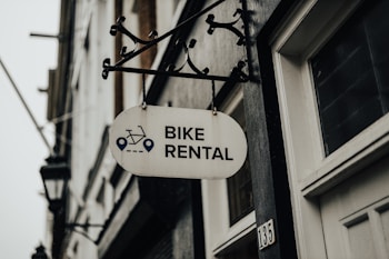 A metal sign with the words 'Bike Rental' hangs from a decorative bracket on a building. The sign includes icons of a bicycle and navigation symbols. The building has a white door and the number '135' visible. The atmosphere appears overcast, contributing to a muted tone.