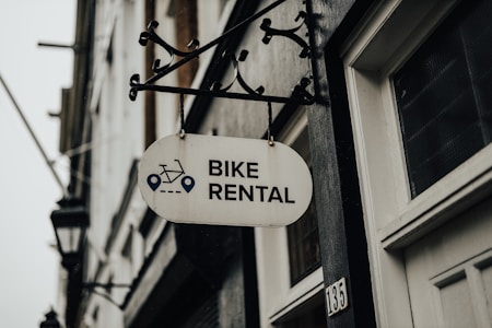 A metal sign with the words 'Bike Rental' hangs from a decorative bracket on a building. The sign includes icons of a bicycle and navigation symbols. The building has a white door and the number '135' visible. The atmosphere appears overcast, contributing to a muted tone.
