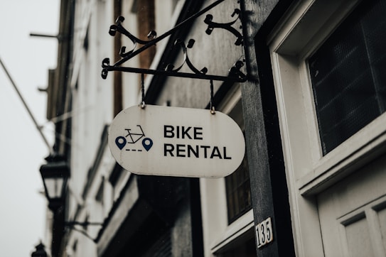 A metal sign with the words 'Bike Rental' hangs from a decorative bracket on a building. The sign includes icons of a bicycle and navigation symbols. The building has a white door and the number '135' visible. The atmosphere appears overcast, contributing to a muted tone.
