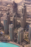 An aerial view of a modern urban landscape featuring several high-rise buildings with intricate architectural designs. The buildings are surrounded by a network of roads, with a construction site visible in the background, indicating ongoing urban development. A large pool or body of water with a bright turquoise color lies in the foreground, adding an element of tranquility to the bustling cityscape.
