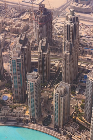 An aerial view of a modern urban landscape featuring several high-rise buildings with intricate architectural designs. The buildings are surrounded by a network of roads, with a construction site visible in the background, indicating ongoing urban development. A large pool or body of water with a bright turquoise color lies in the foreground, adding an element of tranquility to the bustling cityscape.