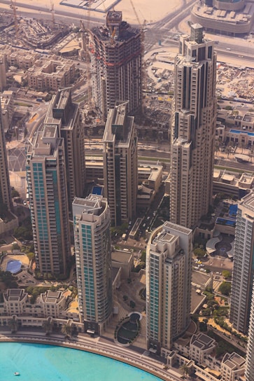 An aerial view of a modern urban landscape featuring several high-rise buildings with intricate architectural designs. The buildings are surrounded by a network of roads, with a construction site visible in the background, indicating ongoing urban development. A large pool or body of water with a bright turquoise color lies in the foreground, adding an element of tranquility to the bustling cityscape.