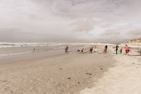 A sandy beach scene with a group of children and a few adults preparing surfboards. The children, dressed in black wetsuits and red tops, appear to be getting ready for surfing lessons. The ocean waves are rolling in, and the sky is overcast with clouds.