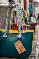 Several colorful handbags with rope handles hang from a metal bar in an outdoor market setting. Each bag has a tag displaying pricing information. The cobblestone path adds a rustic feel.