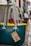 Several colorful handbags with rope handles hang from a metal bar in an outdoor market setting. Each bag has a tag displaying pricing information. The cobblestone path adds a rustic feel.