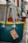 Several colorful handbags with rope handles hang from a metal bar in an outdoor market setting. Each bag has a tag displaying pricing information. The cobblestone path adds a rustic feel.
