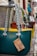 Several colorful handbags with rope handles hang from a metal bar in an outdoor market setting. Each bag has a tag displaying pricing information. The cobblestone path adds a rustic feel.