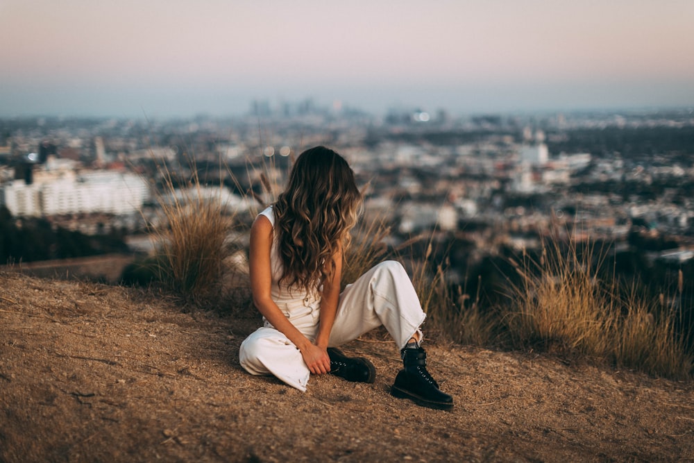 woman wearing white top and pants sitting beside bushes