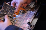 A person is holding a circuit board above an open laptop. The image captures the intricate details of the computer's components, including the motherboard and various connectors.