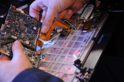 A person is holding a circuit board above an open laptop. The image captures the intricate details of the computer's components, including the motherboard and various connectors.