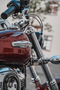 A close-up view of a Harley-Davidson motorcycle, focusing on the handlebar and fuel tank. The chrome details and deep red paint are prominent, with reflections visible on the surface. The background is blurred, highlighting the motorcycle's features.