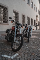 Two parked motorcycles with shiny chrome and black frames are positioned on a cobblestone street. The motorcycles have detailed wheel spokes and handlebars, set against the backdrop of a building with ornate window grills and multiple windows.