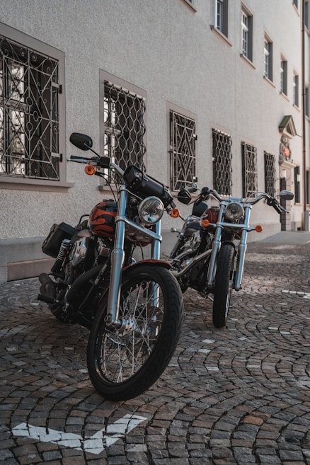 Two parked motorcycles with shiny chrome and black frames are positioned on a cobblestone street. The motorcycles have detailed wheel spokes and handlebars, set against the backdrop of a building with ornate window grills and multiple windows.