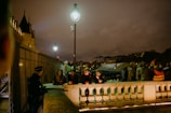 A nighttime scene on a street with a group of people gathered. Some individuals are leaning on a stone railing, with a policeman nearby. A couple of street lamps illuminate the area, which includes a large tent structure in the background, and buildings under a cloudy sky.