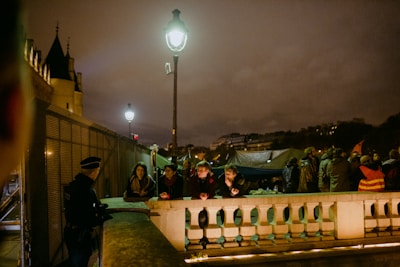 A nighttime scene on a street with a group of people gathered. Some individuals are leaning on a stone railing, with a policeman nearby. A couple of street lamps illuminate the area, which includes a large tent structure in the background, and buildings under a cloudy sky.