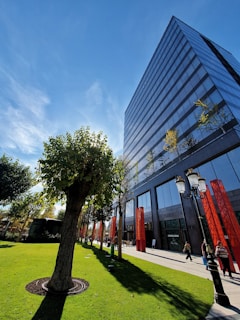 A modern glass office building stands tall under a clear blue sky, reflecting light and partial clouds. In the foreground, a well-maintained lawn with neatly trimmed trees and red pillars lines a path where a few people are strolling. A vintage-style lamppost adds a touch of elegance to the scene.